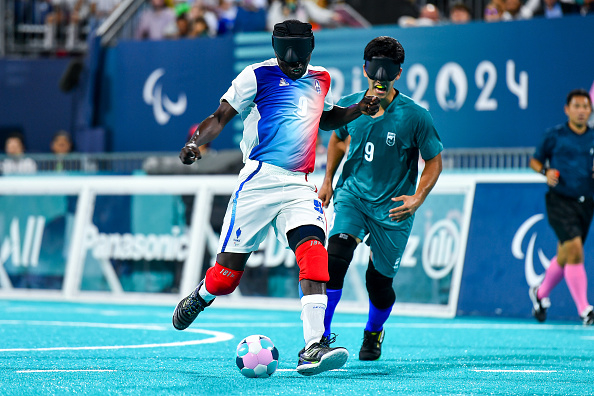 Blind footballers competing in the 2024 Paralympic Games, one in white/red/blue, the other in green, both wearing blindfolds. Referees in the background
