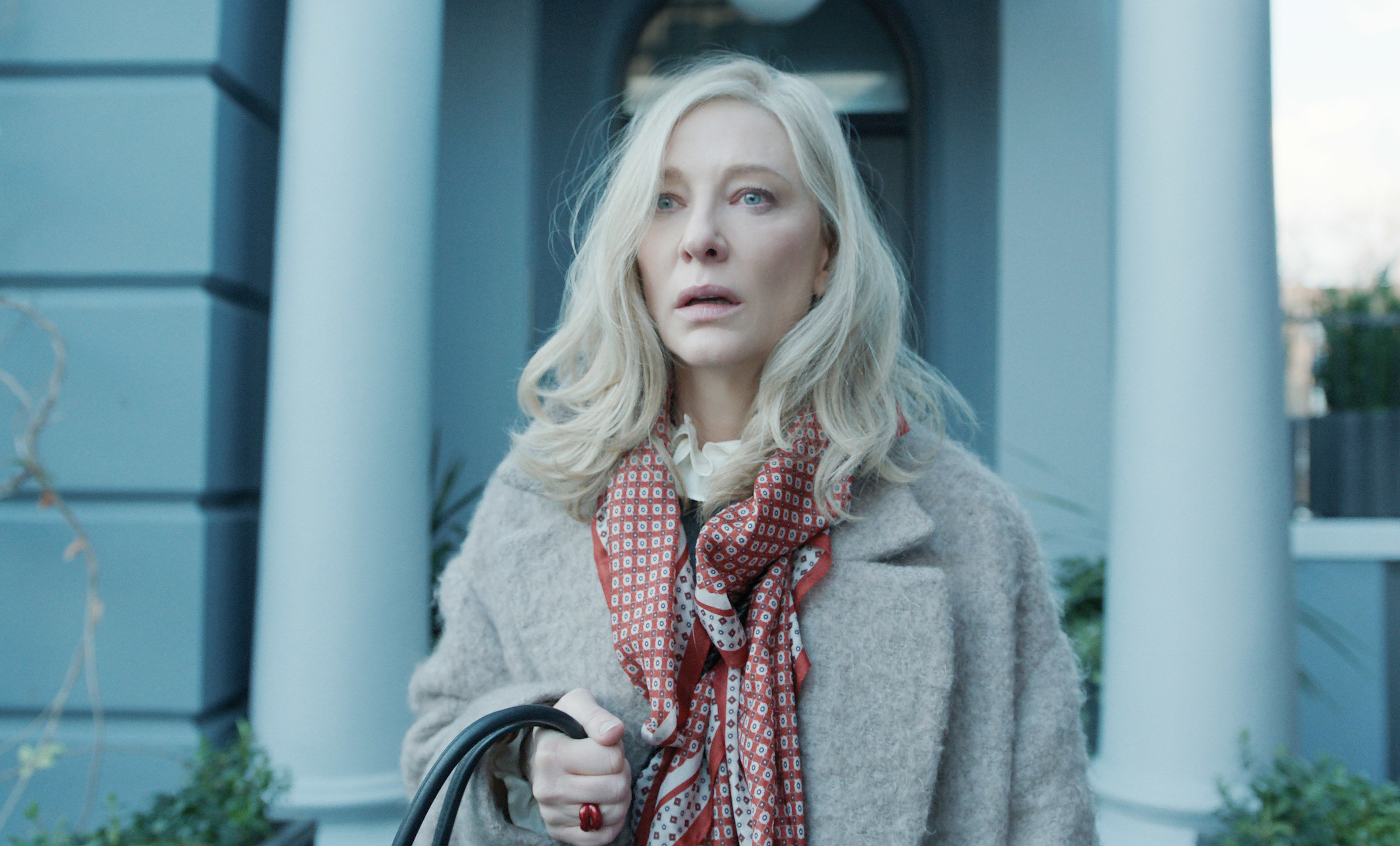 Cate Blanchett in a beige coat and patterned scarf, holding a bag, standing in front of a building with columns