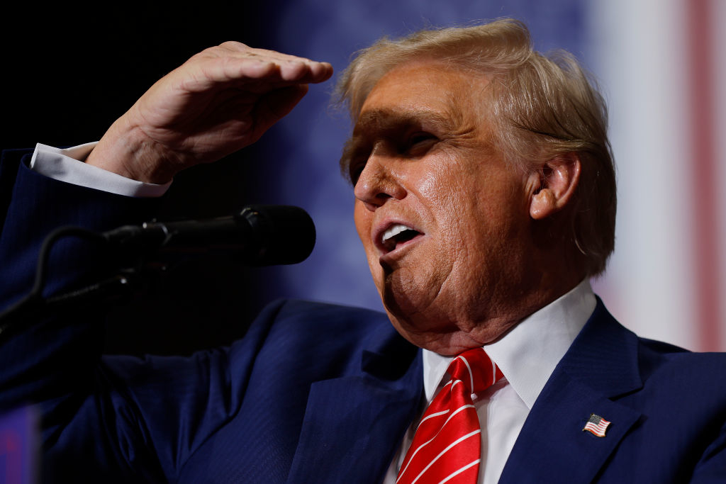 Donald Trump in a suit and tie, holding his hand to his forehead while speaking into a microphone