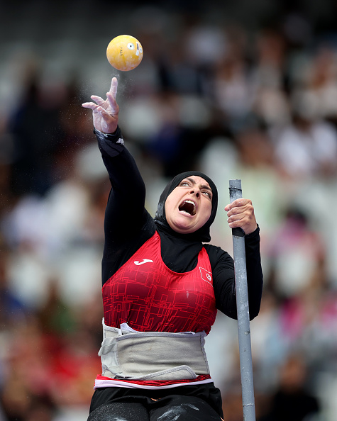 Para-athlete throwing a shot put during a competition, wearing a headscarf, black top, red athletic jersey, and a weightlifting belt