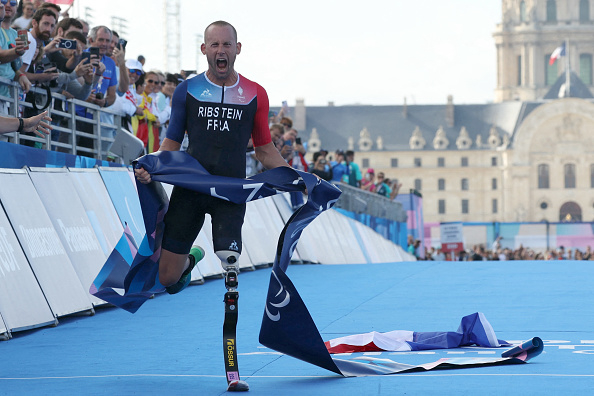 Triathlete Alexis Hanquinquant, dressed in athletic gear, crosses the finish line on a prosthetic leg, cheering with a French flag at a sports event