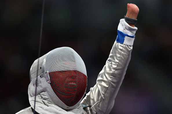 A fencer in full protective gear raises a triumphant fist, displaying a visible disability. The celebratory pose signifies an achievement or victory
