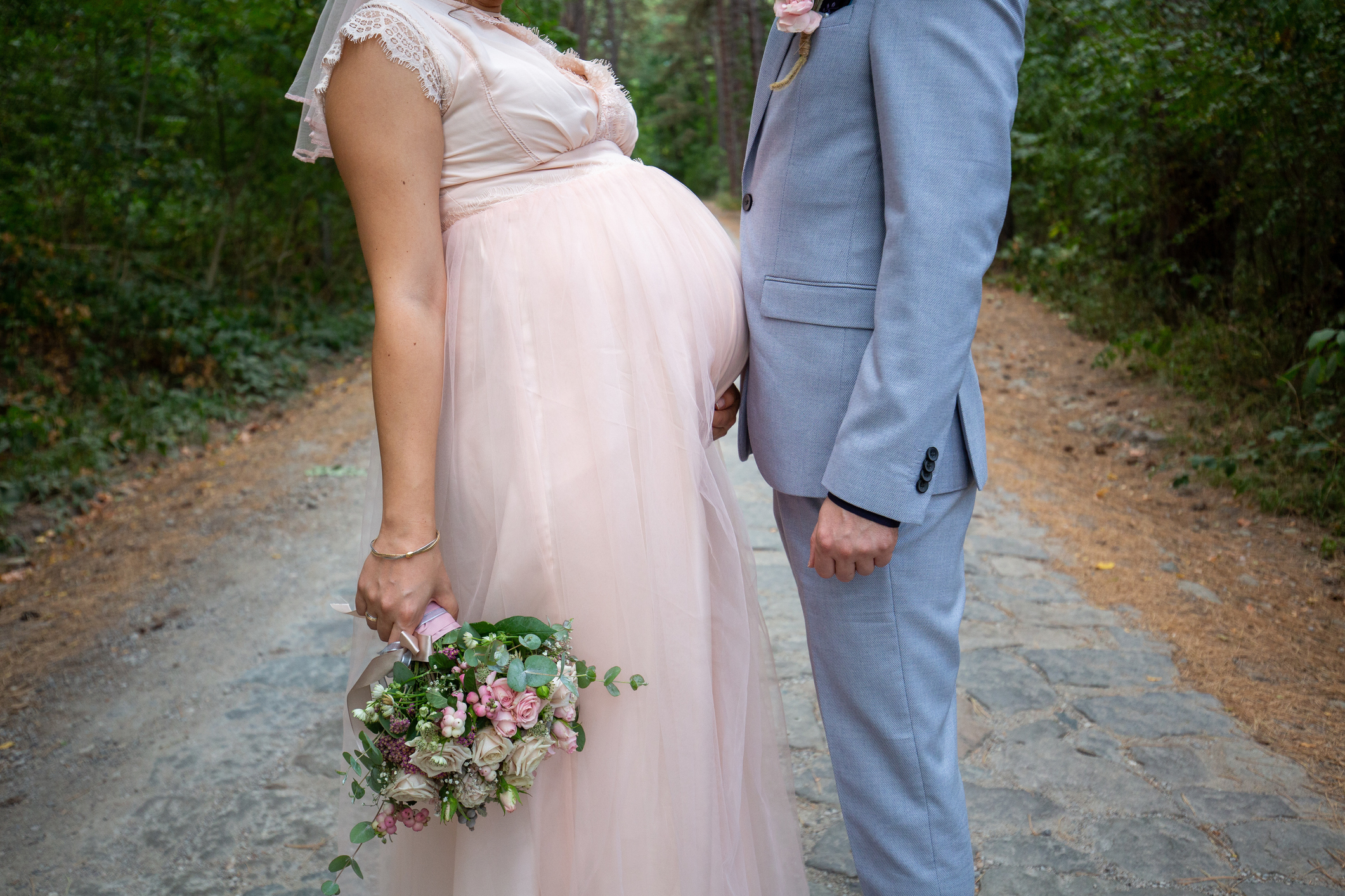 A pregnant bride in a lace-trimmed dress and groom in a light gray suit stand on a rustic path, holding a bouquet of flowers