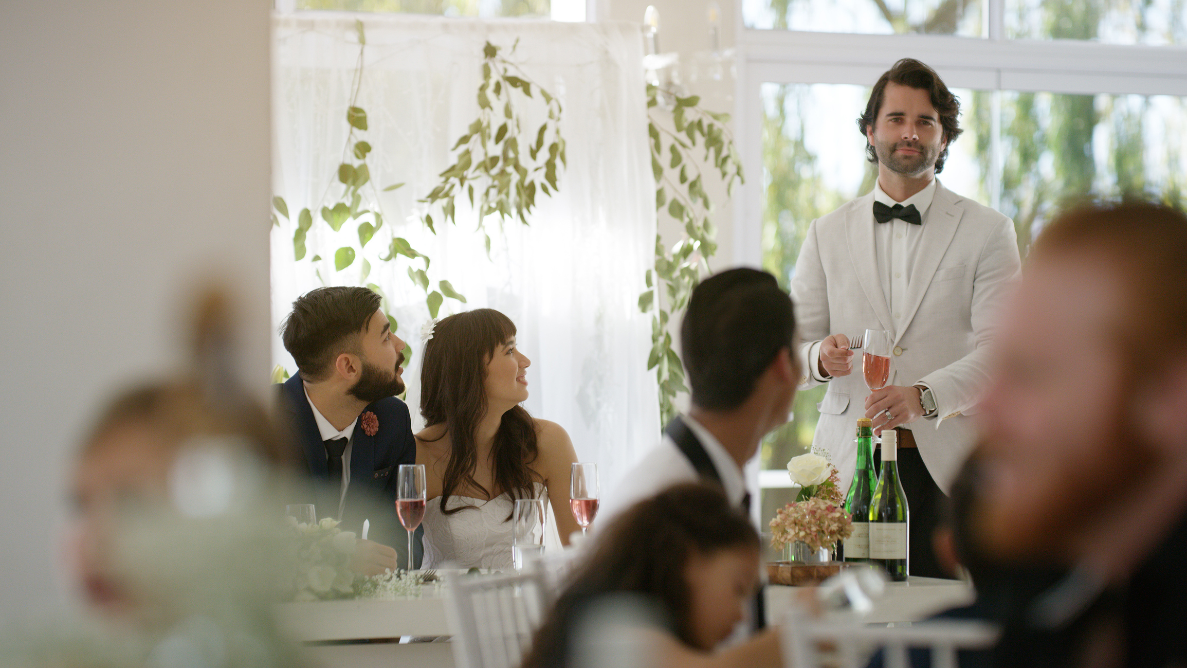 A groom in a white suit gives a speech at his wedding reception, with guests seated and listening attentively