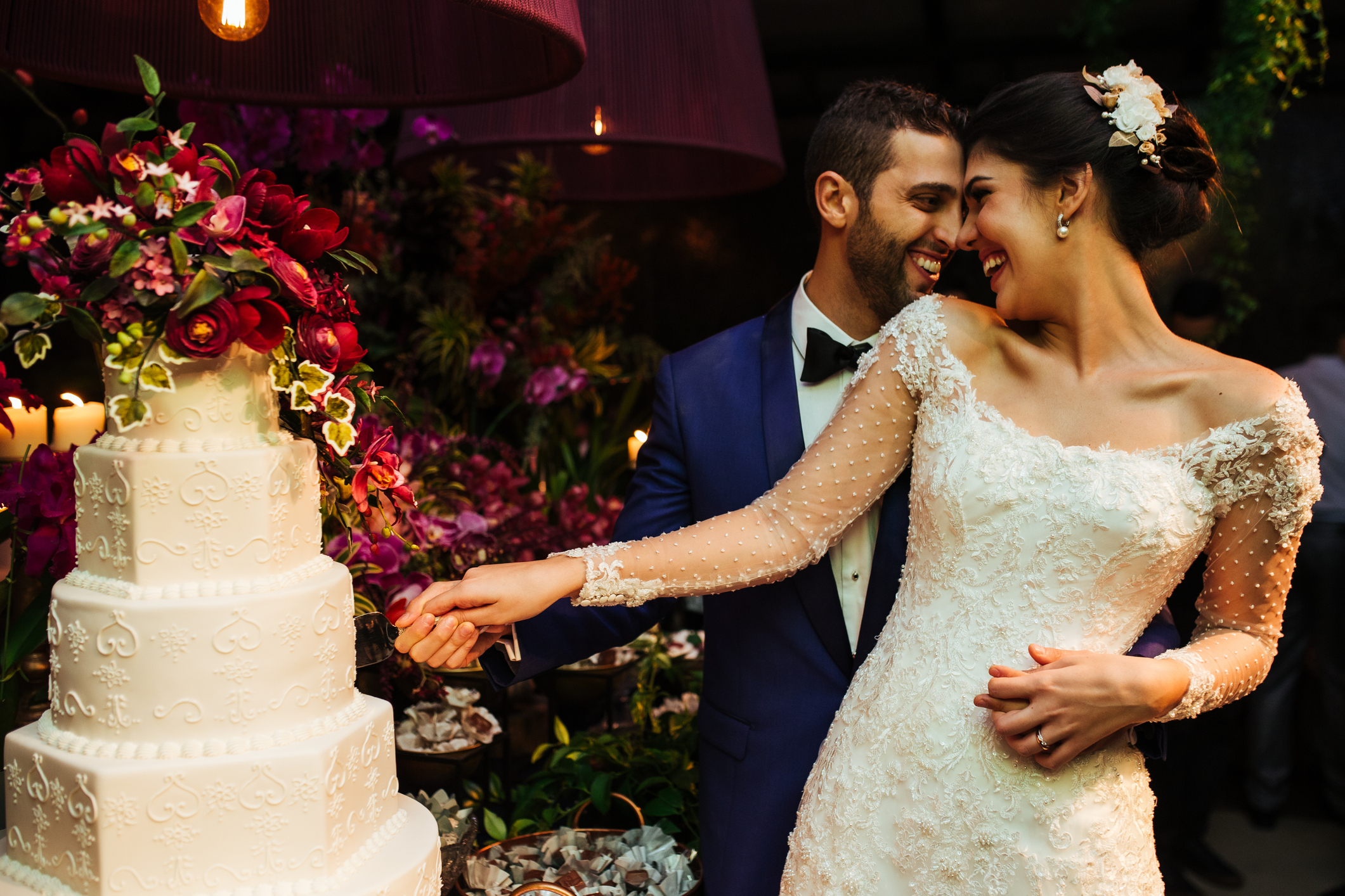 Wedding couple cutting a tiered cake as they smile at each other, wearing formal wedding attire; groom in a tuxedo, bride in a lace gown with floral hairpiece