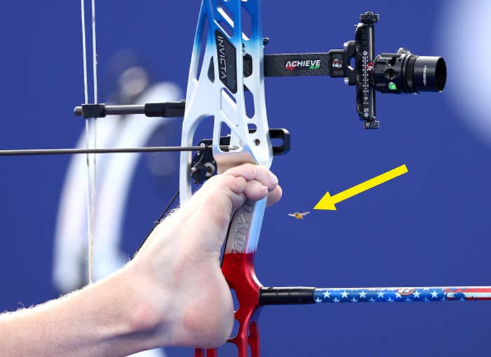 An archer uses his foot to aim a bow, focusing intently on the target at a competitive event
