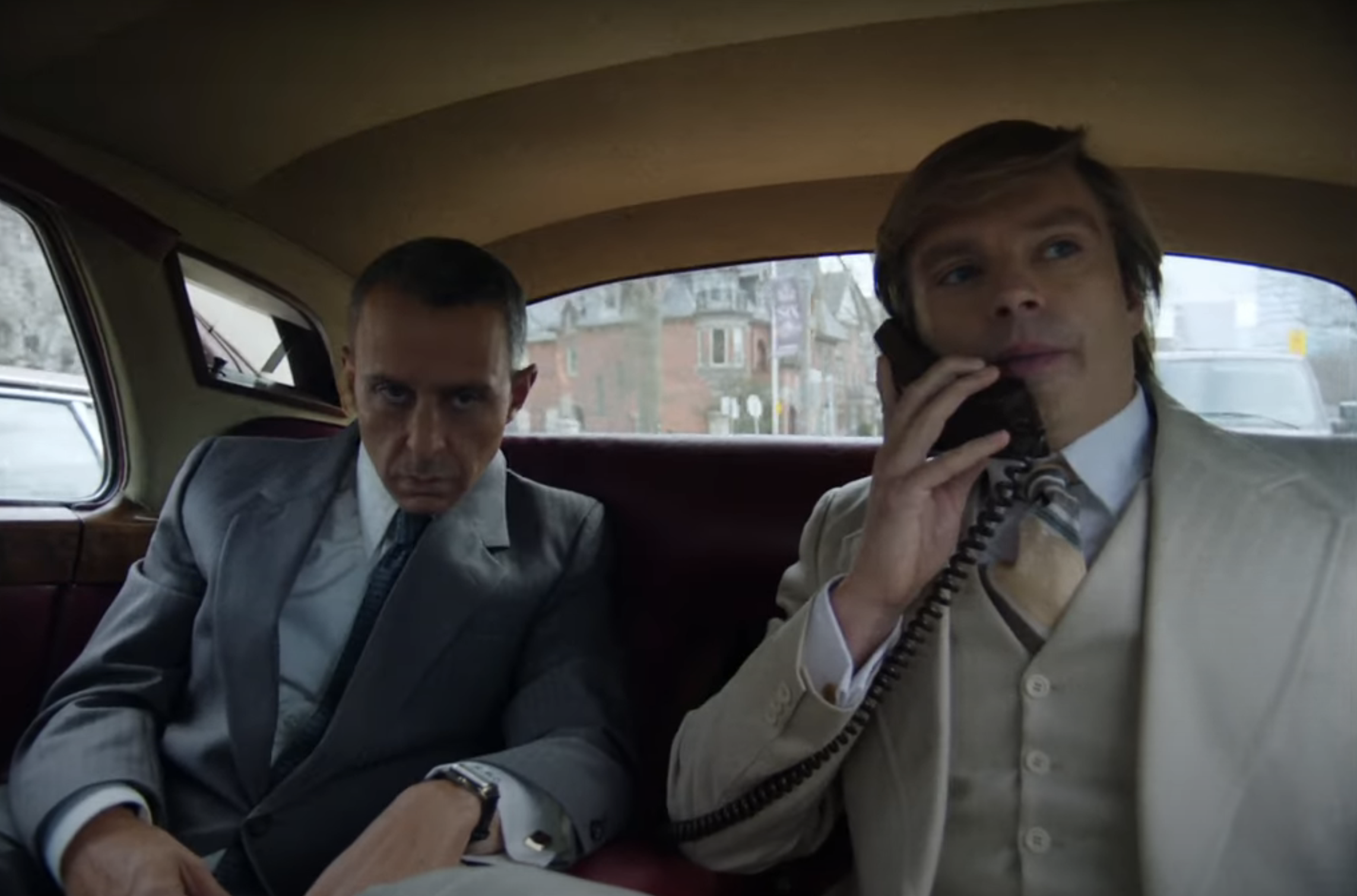 Two men sit in the back of a vintage car. The left man glares while the right man talks on a phone. Both are dressed in formal suits. Names unknown