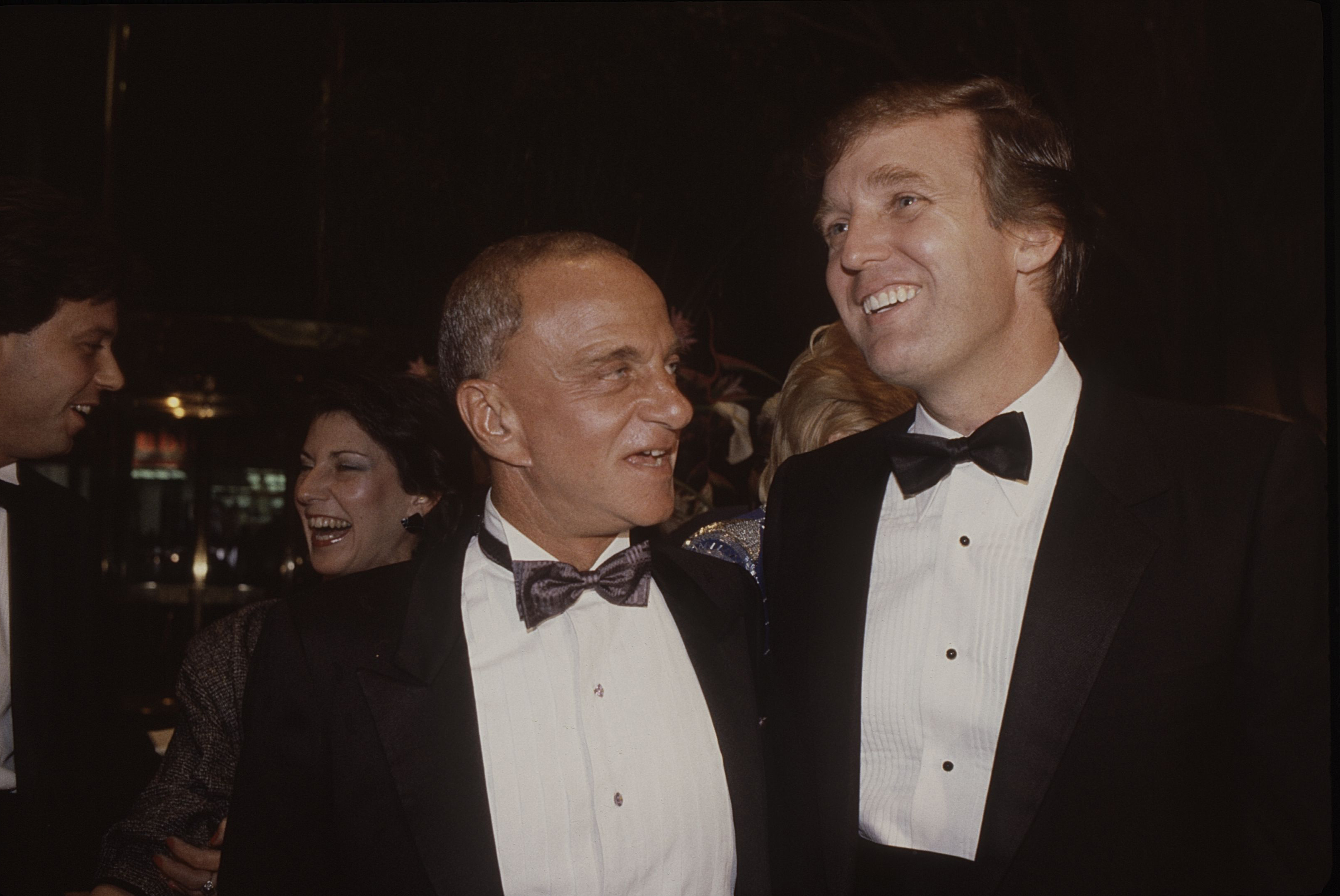 Roy Cohn and Donald Trump smiling and talking at an event, both in tuxedos. Two other people are partially visible in the background