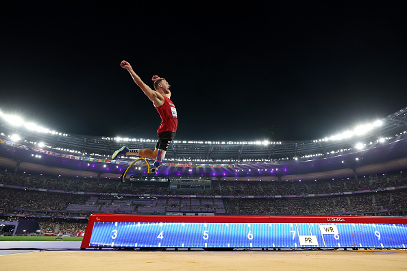 Para-athlete Markus Rehm competes in a long jump event at a stadium, captured mid-air with a dynamic pose and a prosthetic leg visible