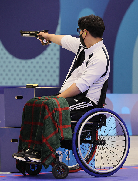 A person in a wheelchair wearing a white sports shirt and black mask aims a pistol during a shooting event, covered with a green and red blanket