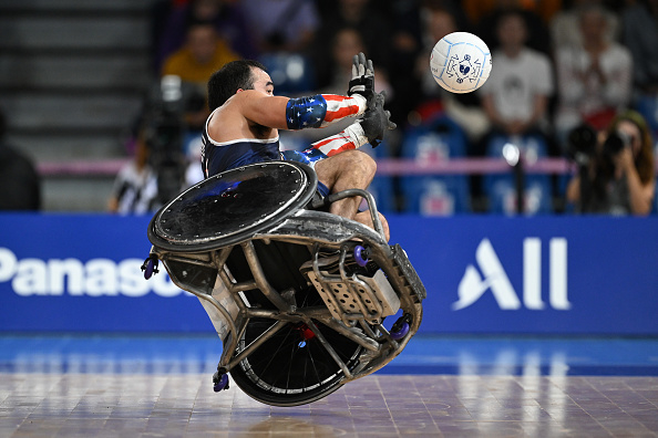 A wheelchair rugby player catches the ball mid-air during a game, showcasing intense action and athleticism. Fans and photographers are seen in the background