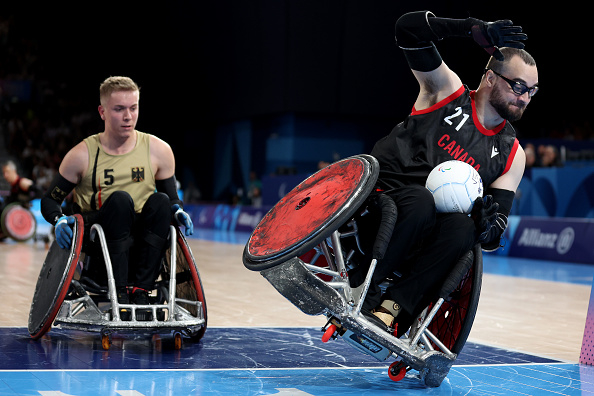Canada's #21 and Germany's #5 compete in a wheelchair rugby match. The Canadian player, holding the ball, moves aggressively towards the camera