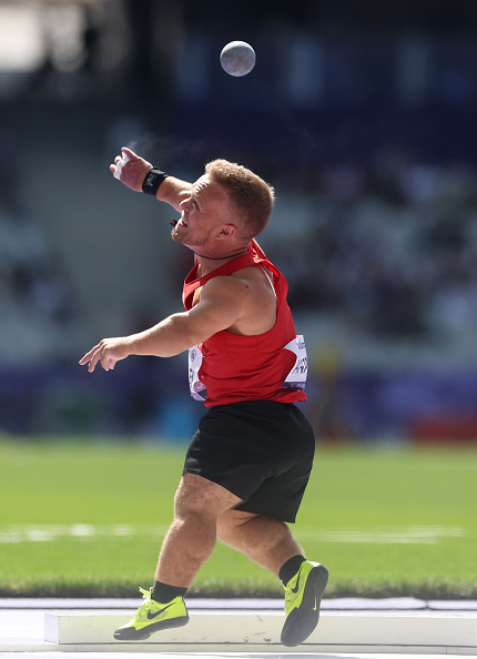 Person participating in a shot put event, wearing a red tank top, black shorts, and neon green shoes, captured mid-throw with the shot in the air