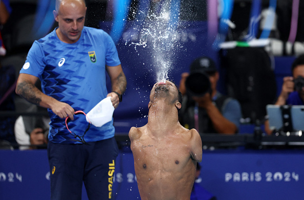 Paralympian Daniel Dias, shirtless, exhales water upward after swimming at the Paris 2024 event. A coach stands nearby holding a cap and mask