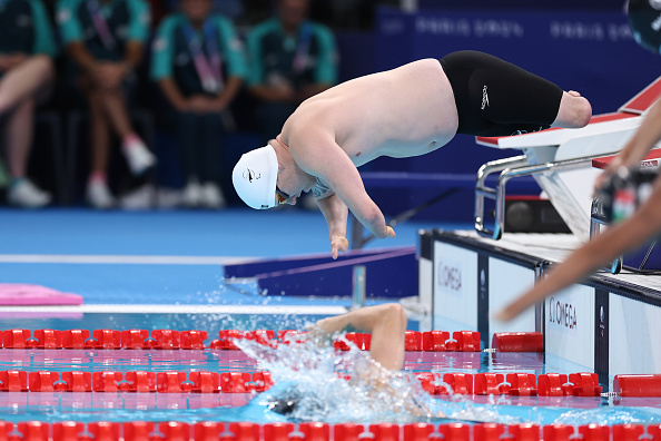 A male swimmer dressed in swimwear and goggles dives into the pool as another swimmer creates a splash, seen mid-race at a competitive swimming event