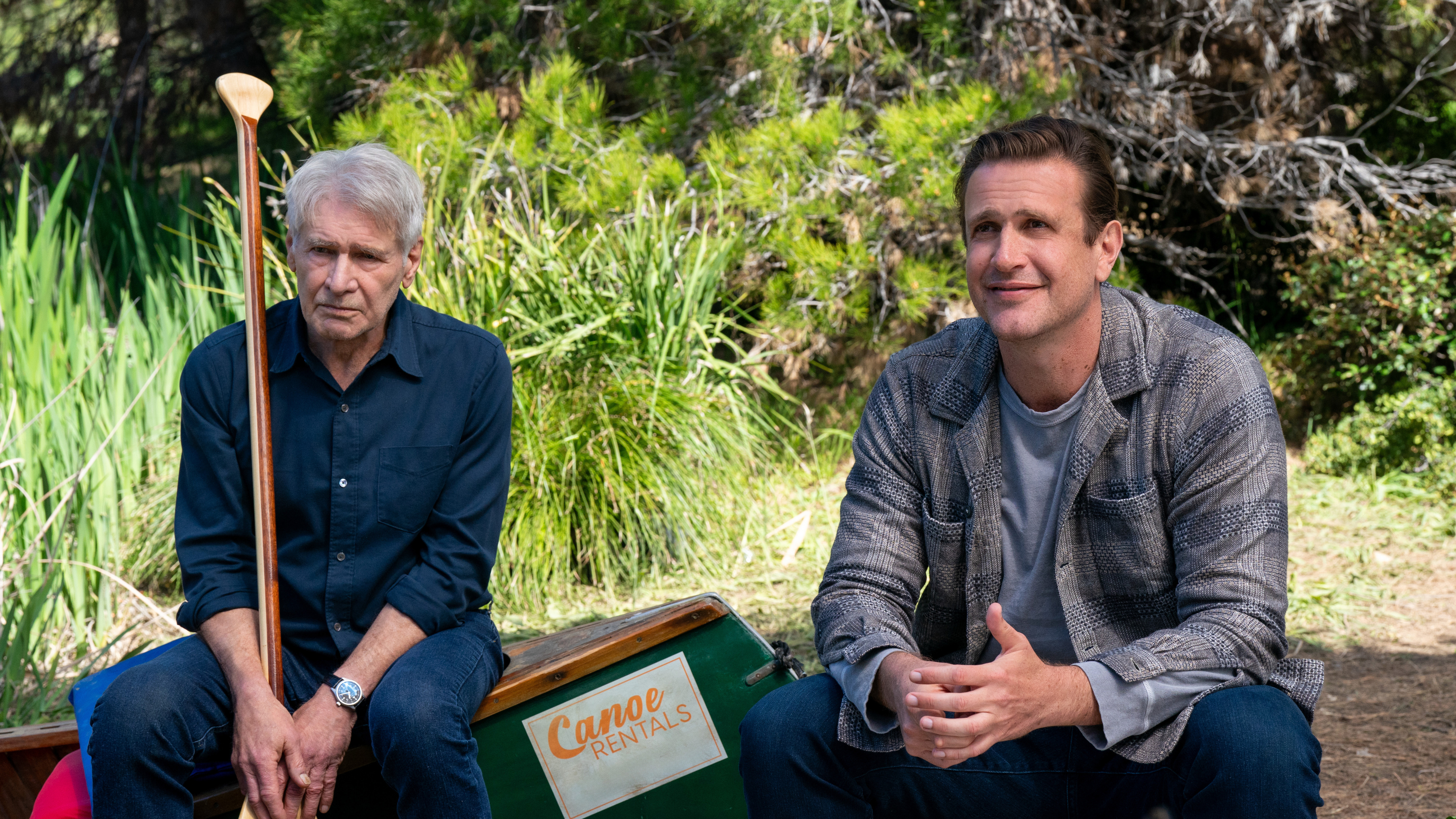 Harrison Ford and Jason Segel sit outdoors next to a canoe labeled "Canoe Rentals." Ford holds a paddle, and Segel appears engaged in conversation