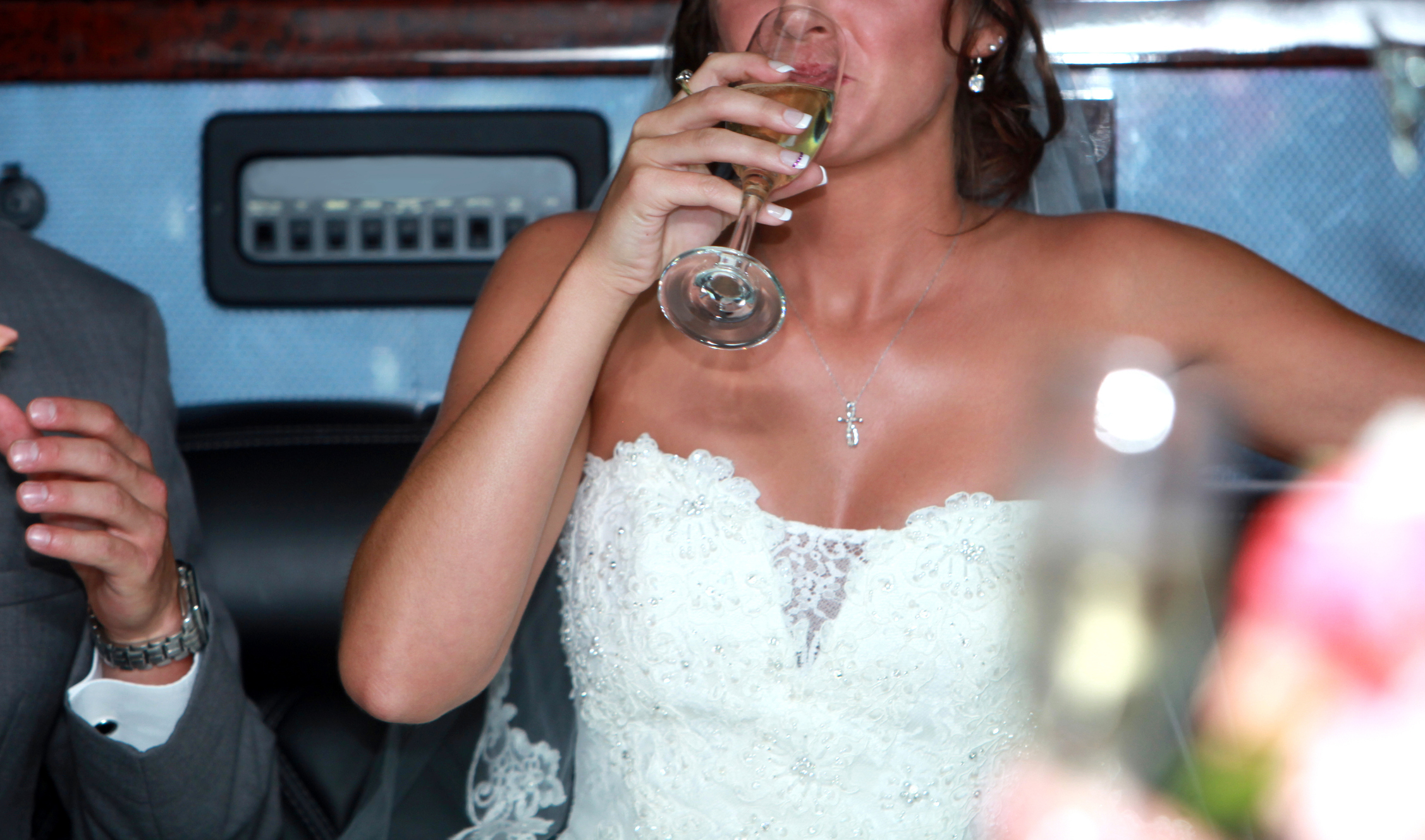 Bride in a strapless lace wedding dress sips champagne, seated beside a man in a grey suit