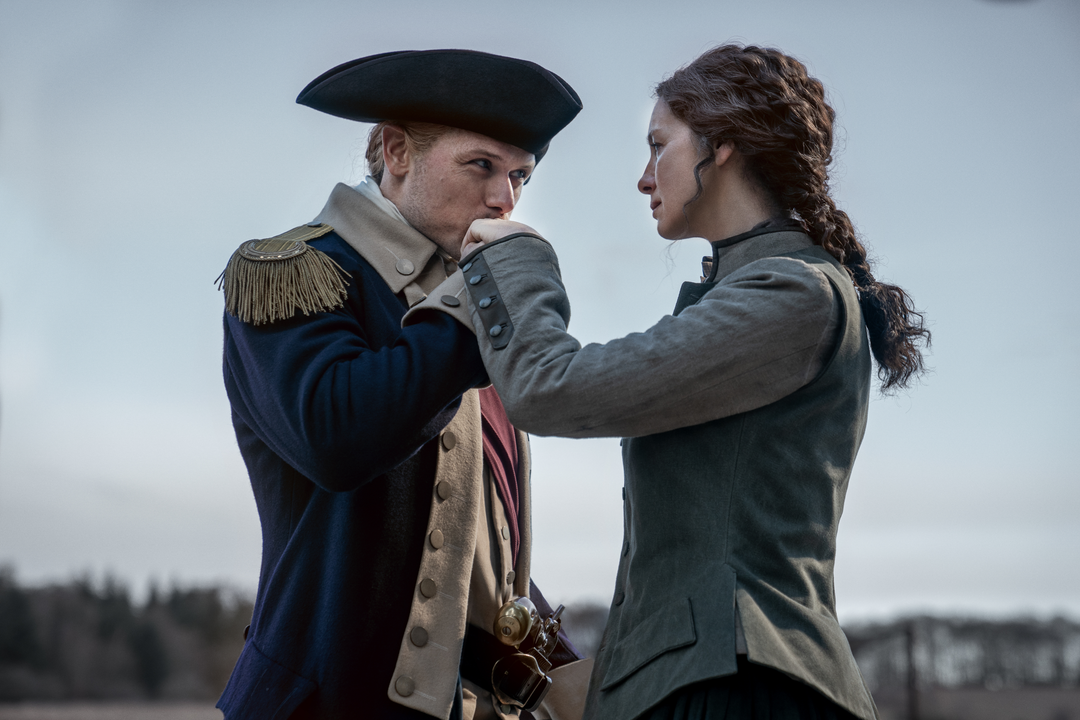 Sam Heughan, in historical military attire, gently kisses the hand of Caitriona Balfe, who is dressed in period clothing, in an outdoor setting