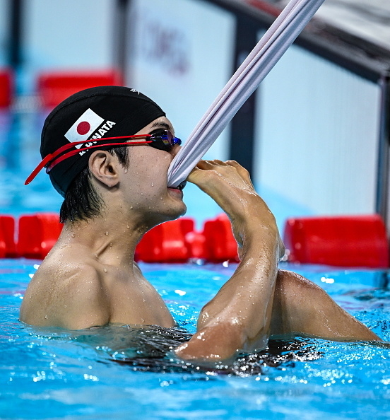 Japanese swimmer in competition pool, biting a towel to remove his swim cap while taking a break at a swimming event