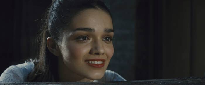 Rachel Zegler smiles while looking slightly upward, head resting on her arms, appearing wistful or hopeful