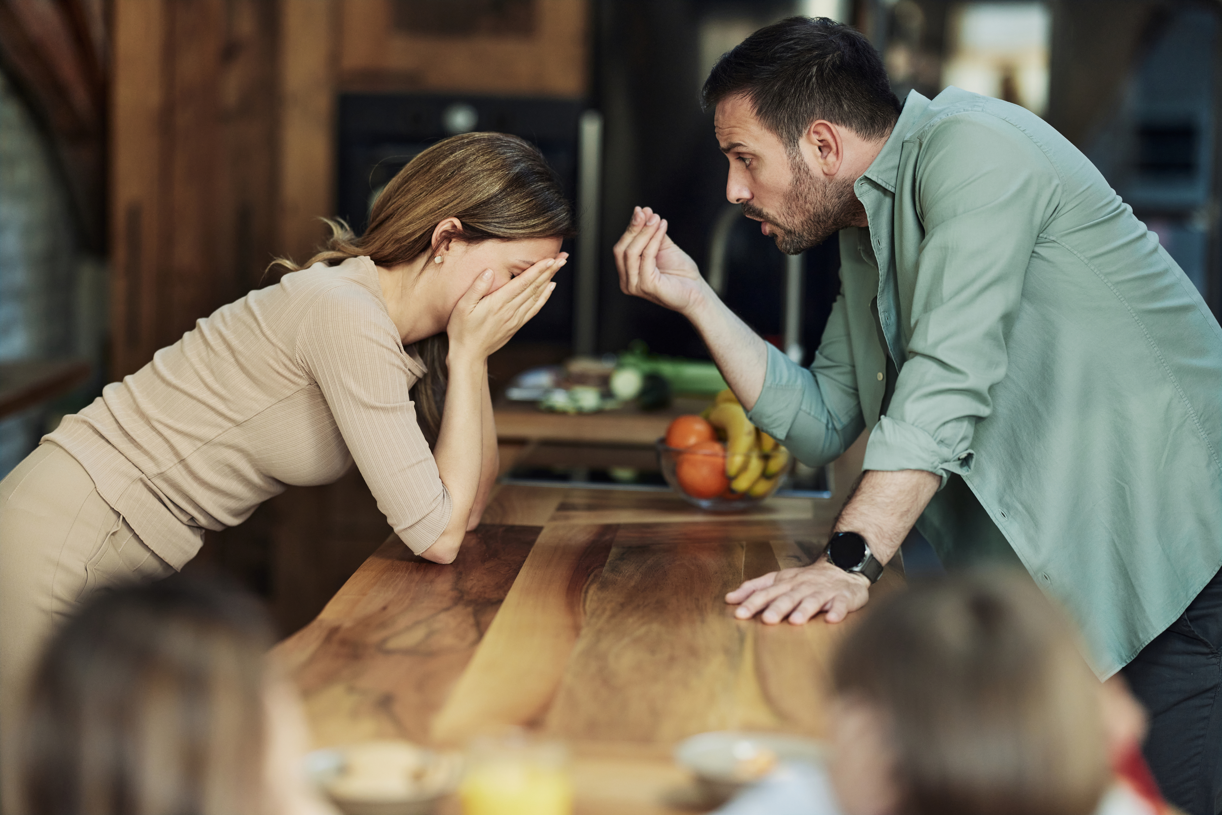 A woman covers her face in distress while a man gestures angrily at her across a kitchen counter. Two children are blurred in the foreground