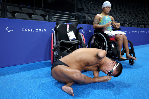 Two Paralympic swimmers, one on the ground stretching and the other in a wheelchair preparing by the pool at the Paris 2024 Paralympics