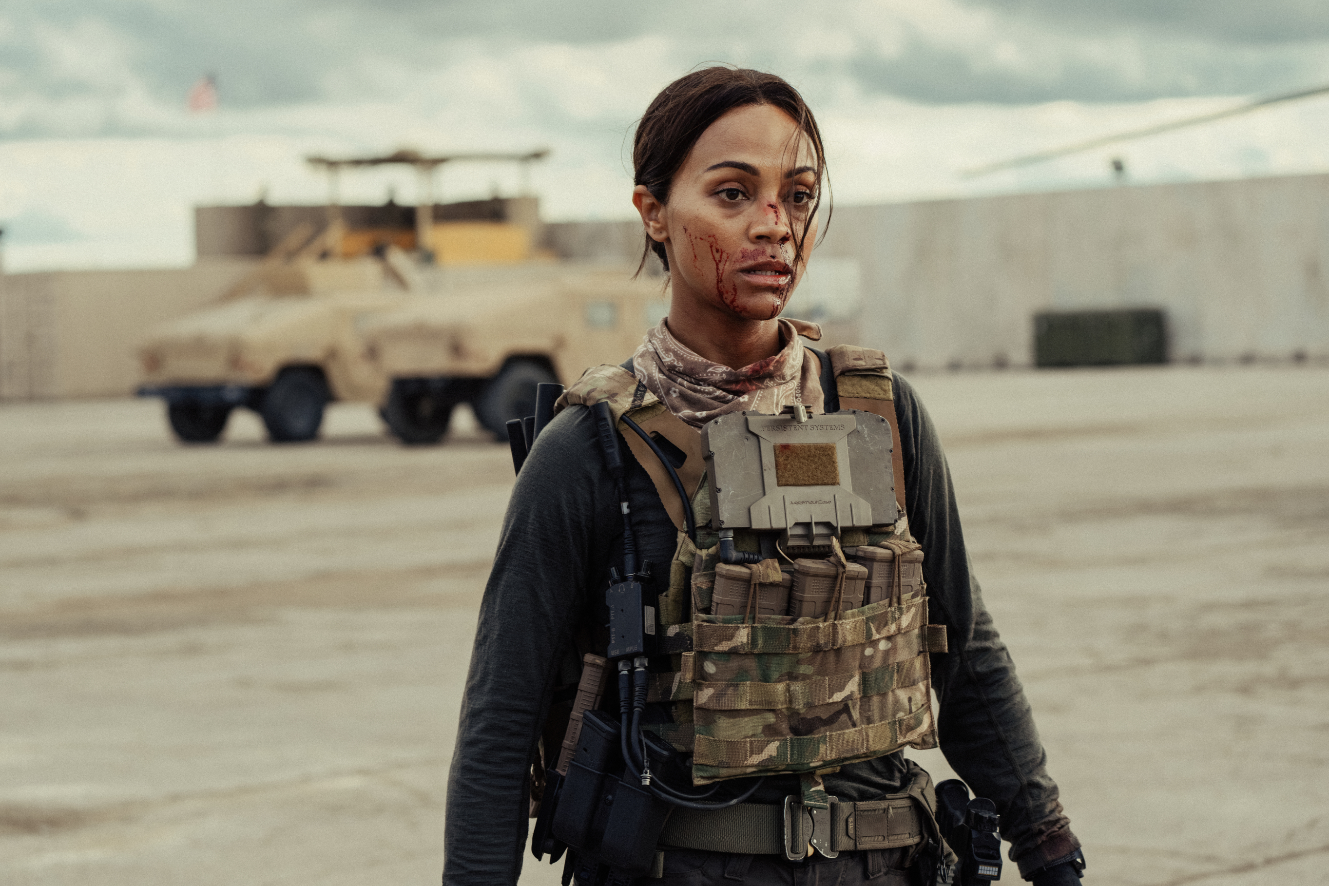 Zoe Saldana in military gear with a tactical vest and camo, on a base with armed vehicles in the background. She has a serious expression and a wound on her face
