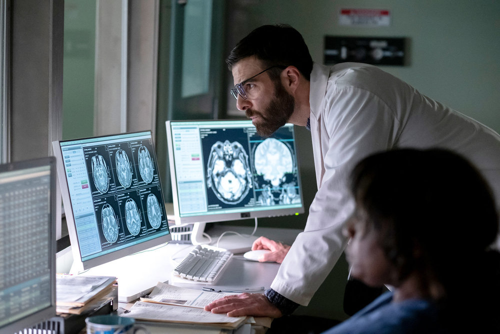 Zachary Quinto observing MRI brain scans on monitors in a medical office, with another person seated nearby