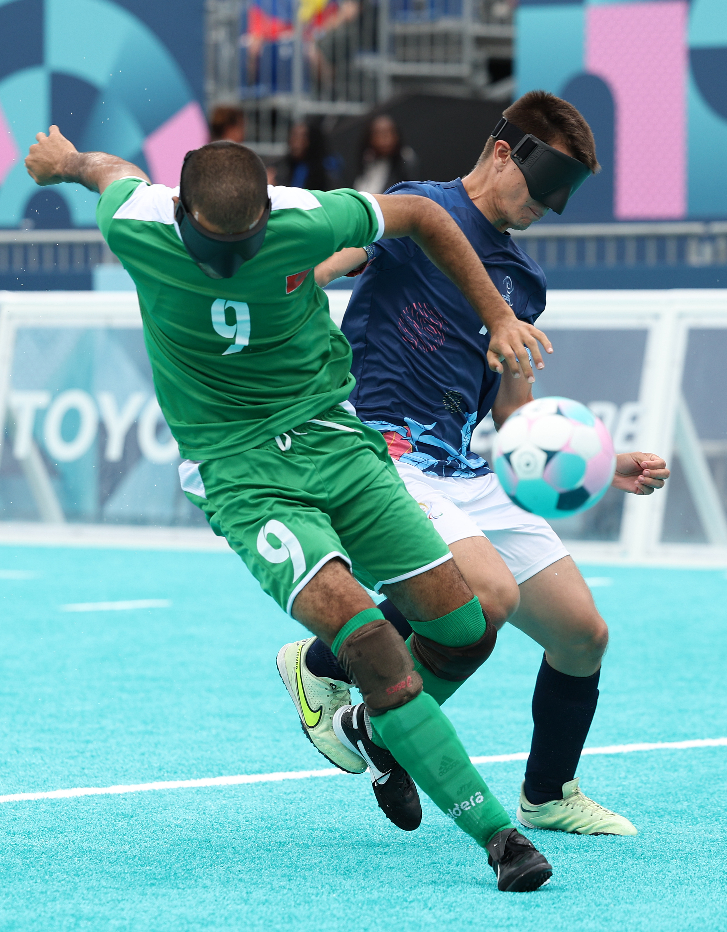 Visually impaired athletes engage in a soccer match, both wearing eye shades. One player in a green uniform appears to be taking a shot, while another in blue defends