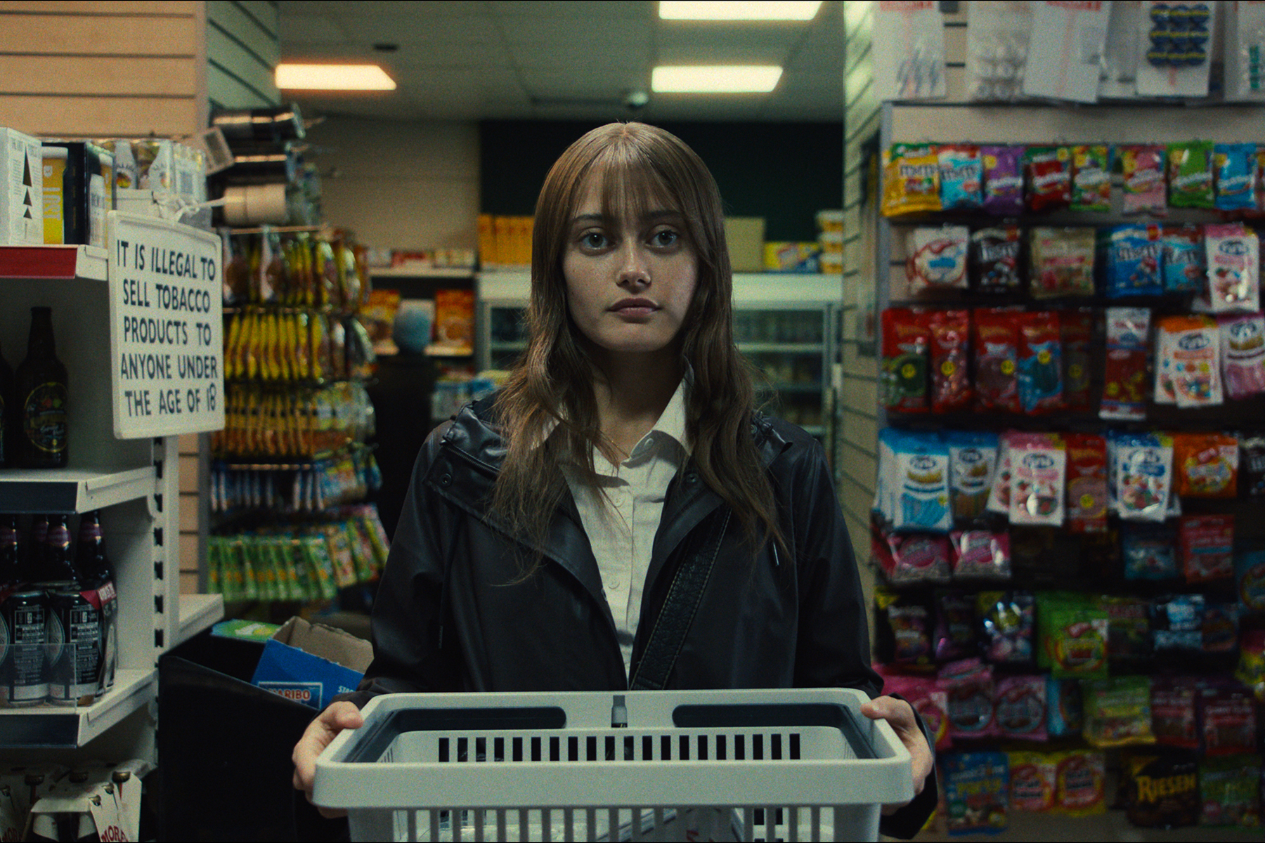 Ella Purnell stands in a convenience store holding a shopping basket, surrounded by packed shelves and a sign stating the legal age for purchasing tobacco products