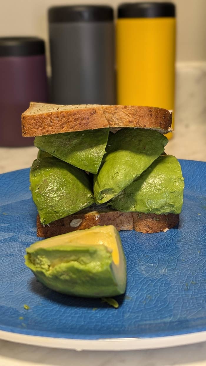A thick sandwich consisting of two slices of bread and several large avocado slices, placed on a blue plate, with three insulated bottles in the background