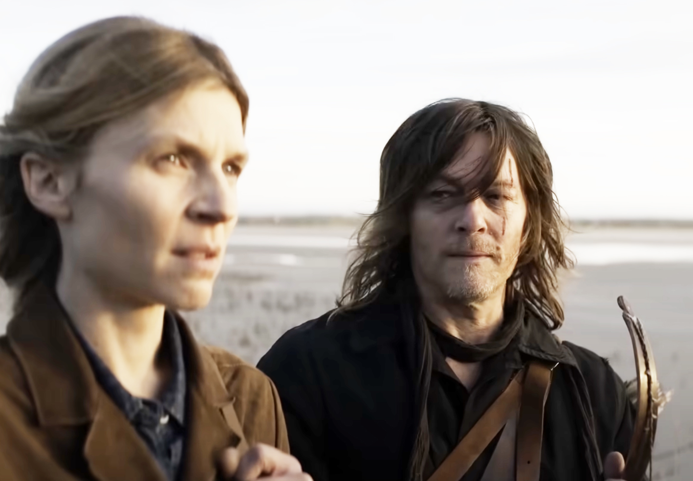 Clémence Poésy and Norman Reedus stand together outdoors, looking into the distance. Poésy is on the left, and Reedus is on the right holding a weapon