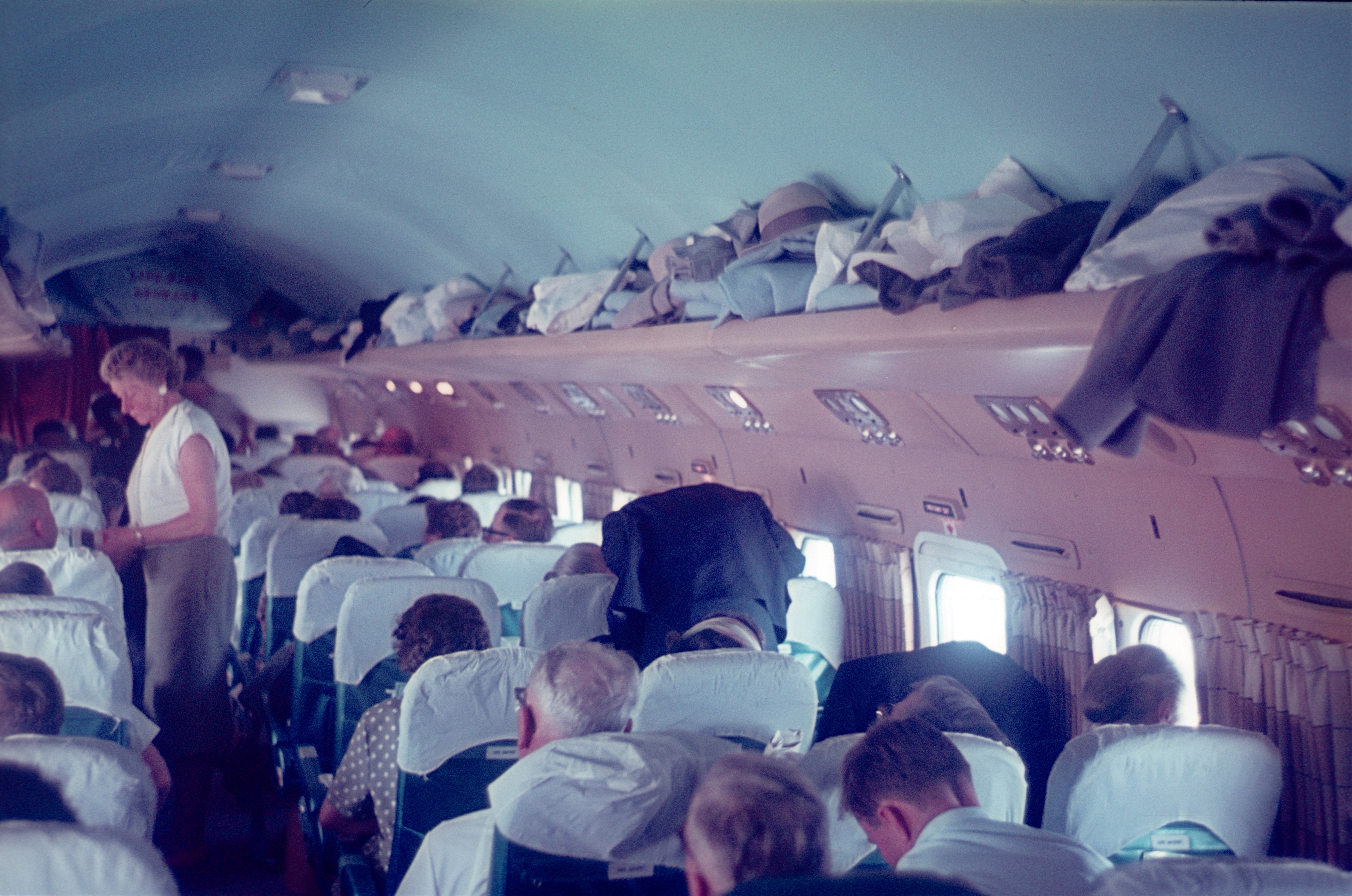 Passengers sit in rows and store luggage in overhead compartments on a crowded airplane. A flight attendant assists in the aisle