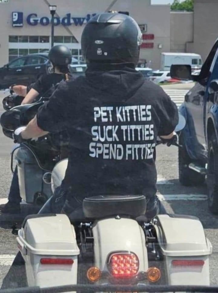 A motorcyclist is wearing a black helmet and T-shirt with the text "Pet kitties, suck titties, spend fitties" on the back. Another biker is seen ahead