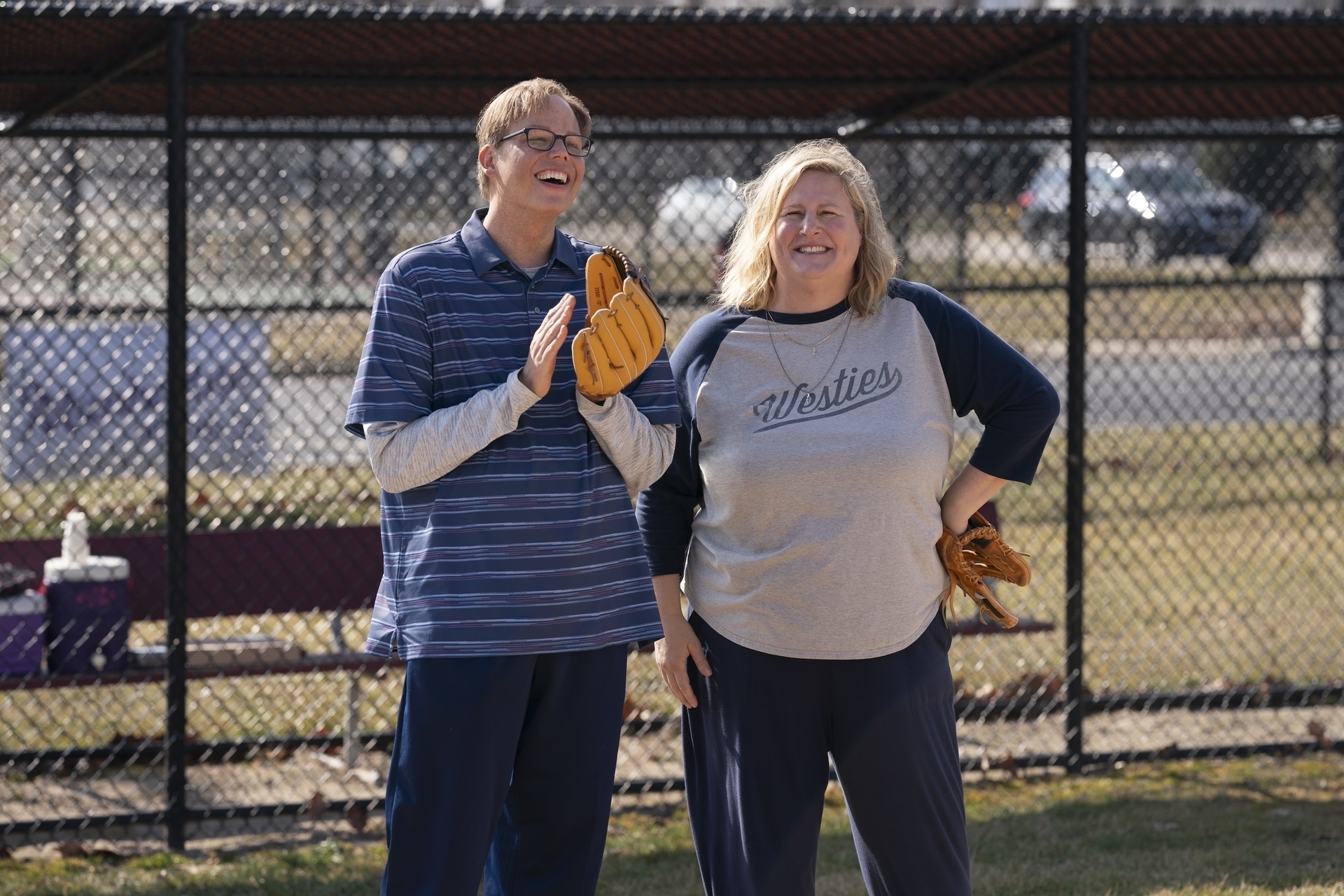 Jeff Hiller and Bridget Everett, dressed casually with baseball gloves, sharing a moment on a baseball field