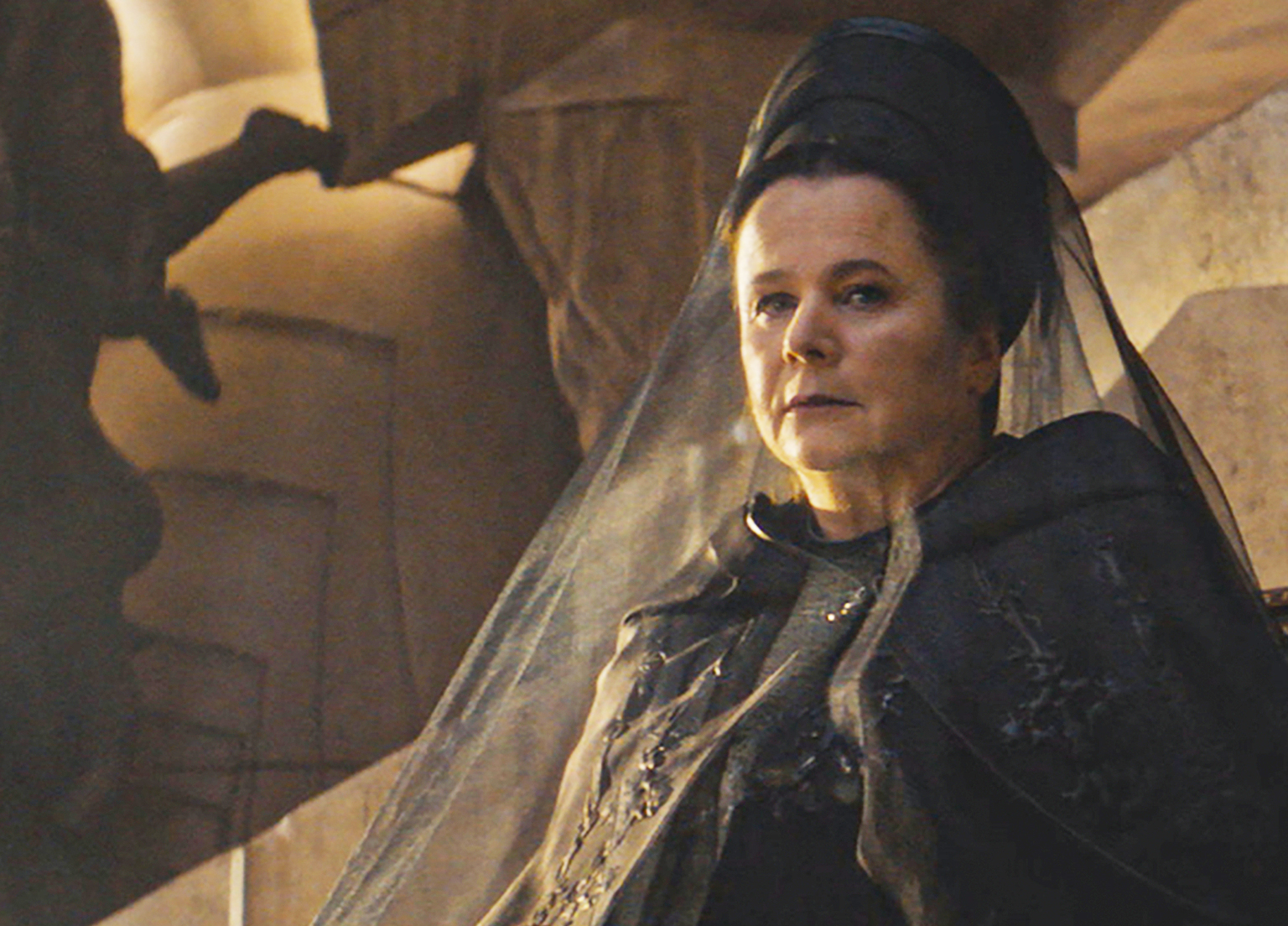 Emily Watson in an ornate, medieval-style gown and headpiece with a sheer veil, appearing serious in a dramatic setting