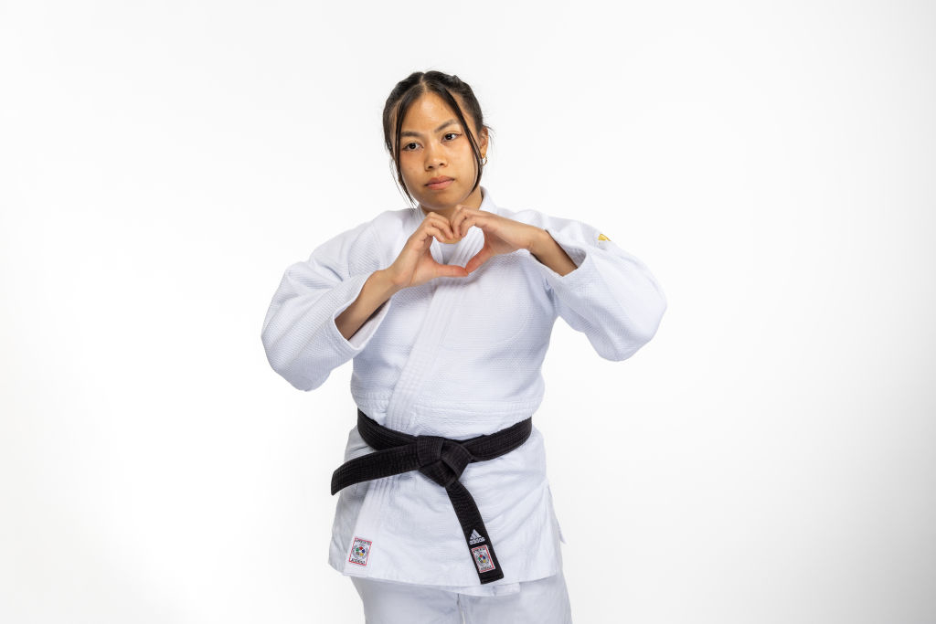 Liana Mutia performs a heart gesture while wearing a judo gi and black belt against a plain background