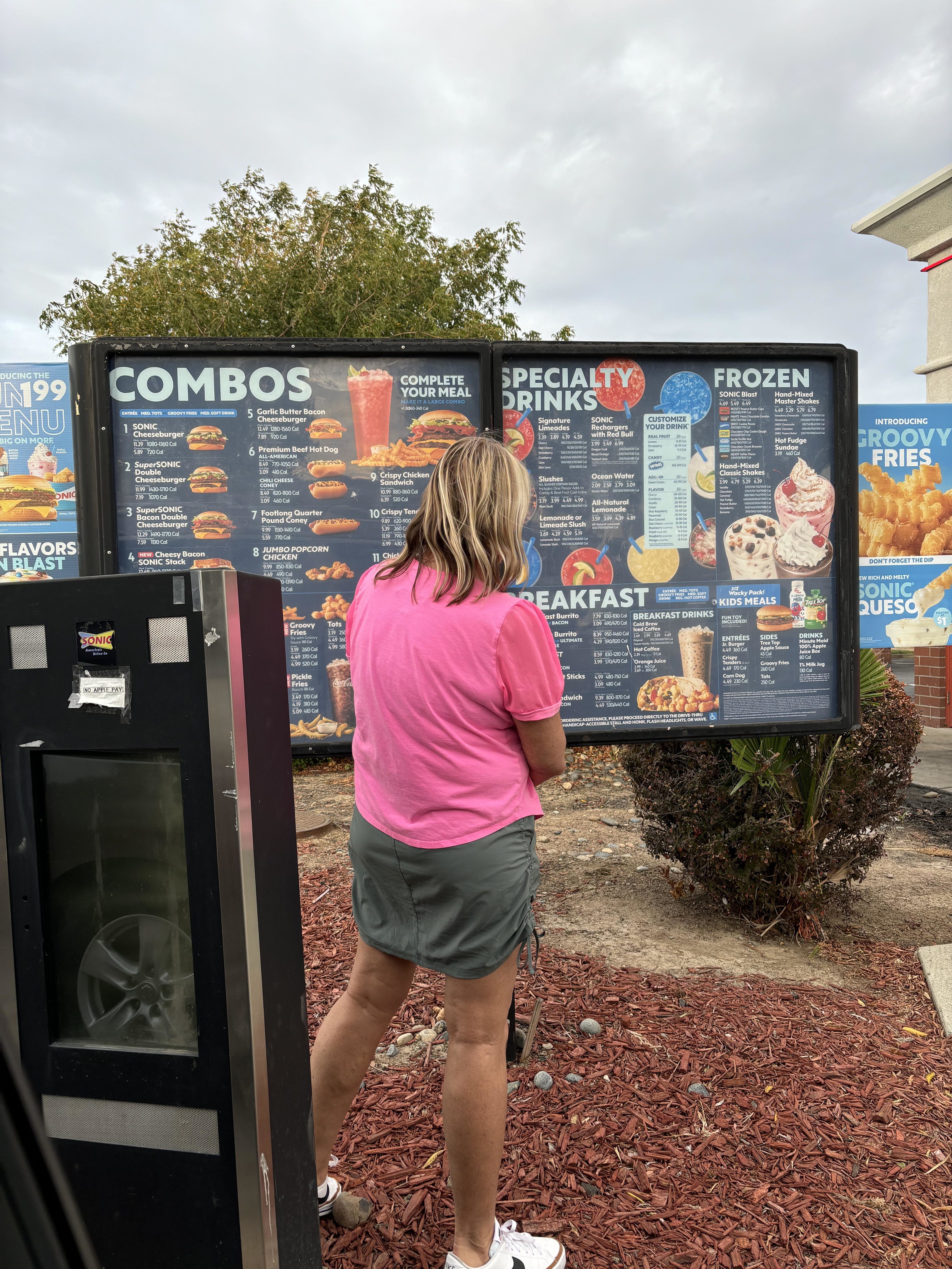 A person stands at a drive-through menu board at a fast-food restaurant, looking at food and drink options