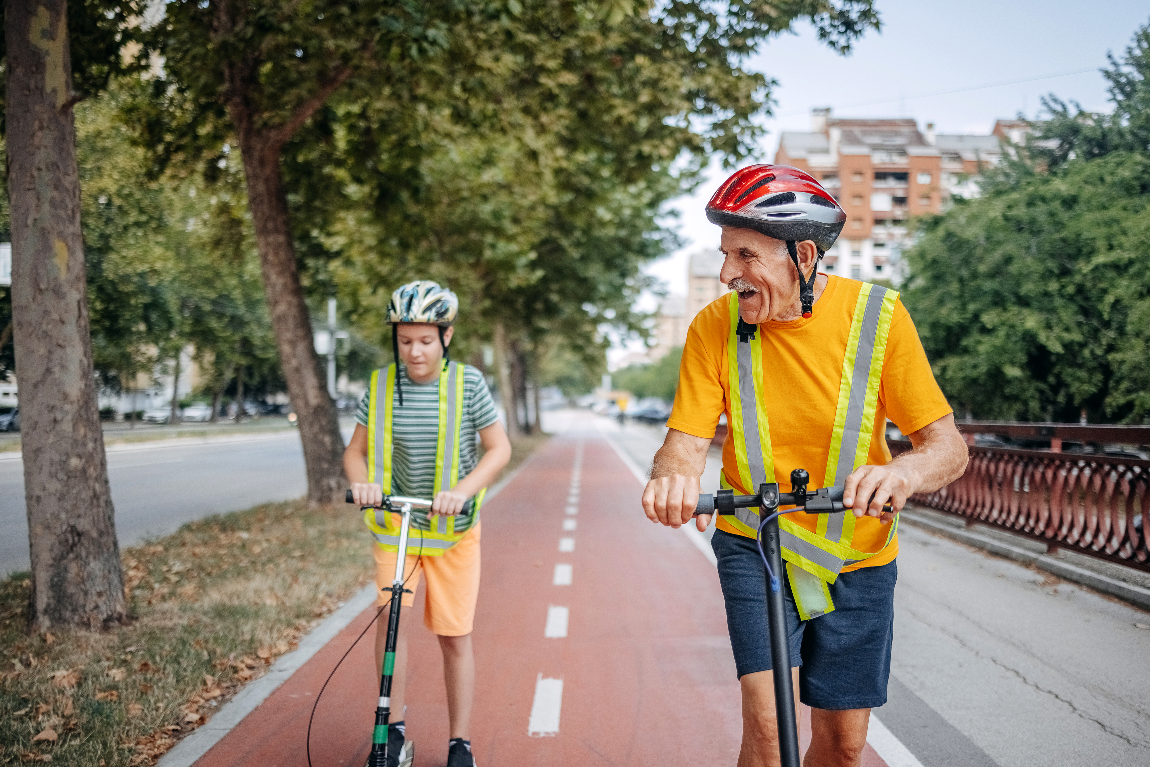 An older man and a young boy ride scooters on a bike path. Both wear helmets and reflective vests for safety. Trees and buildings are visible in the background