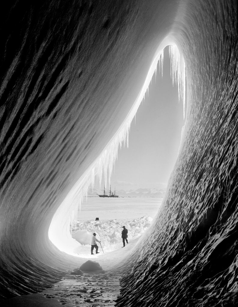 Two individuals stand at the entrance of an ice cave with icicles hanging from the ceiling, while a ship is seen in the snowy landscape outside