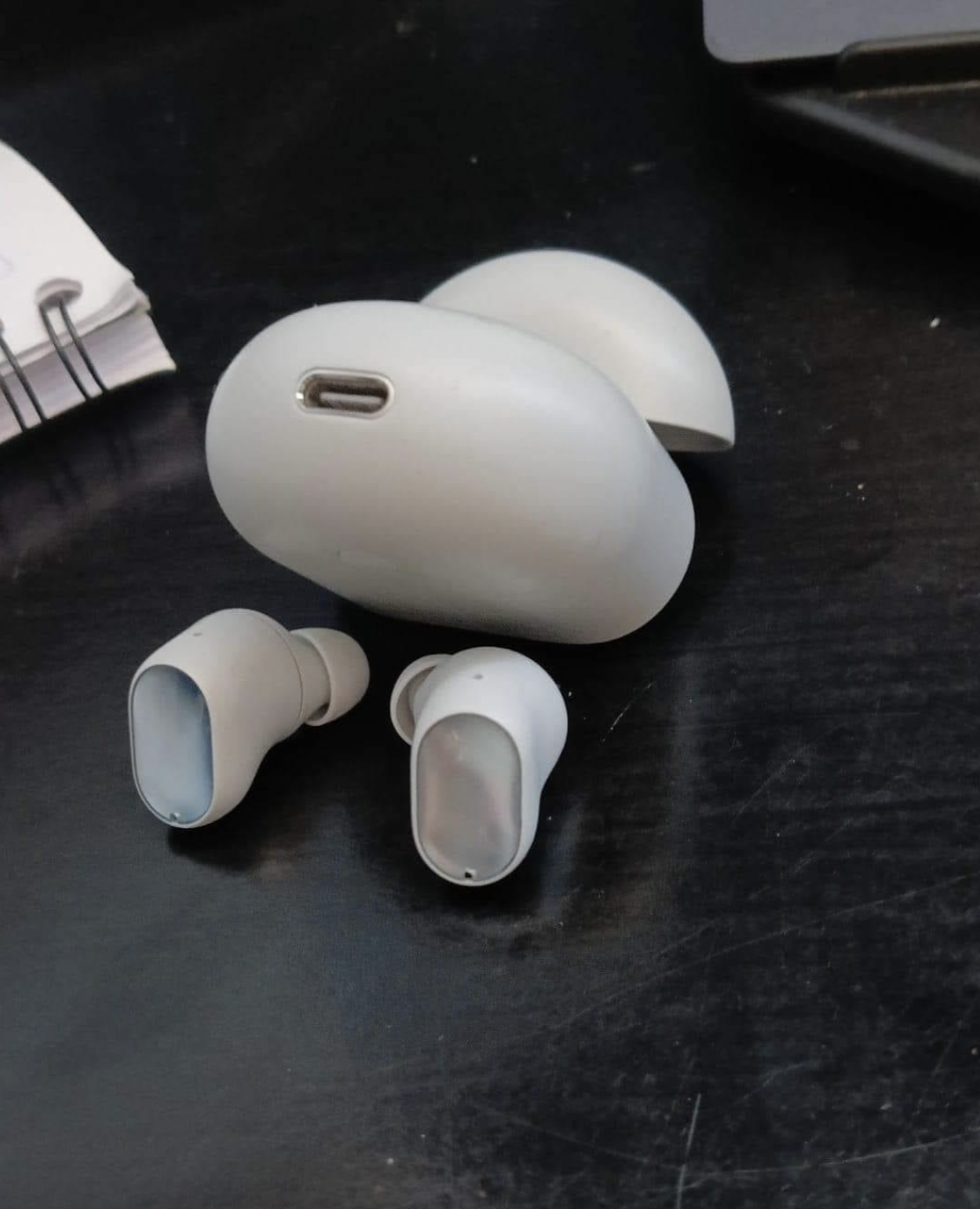 White wireless earbuds with charging case on a desk beside a notebook and laptop