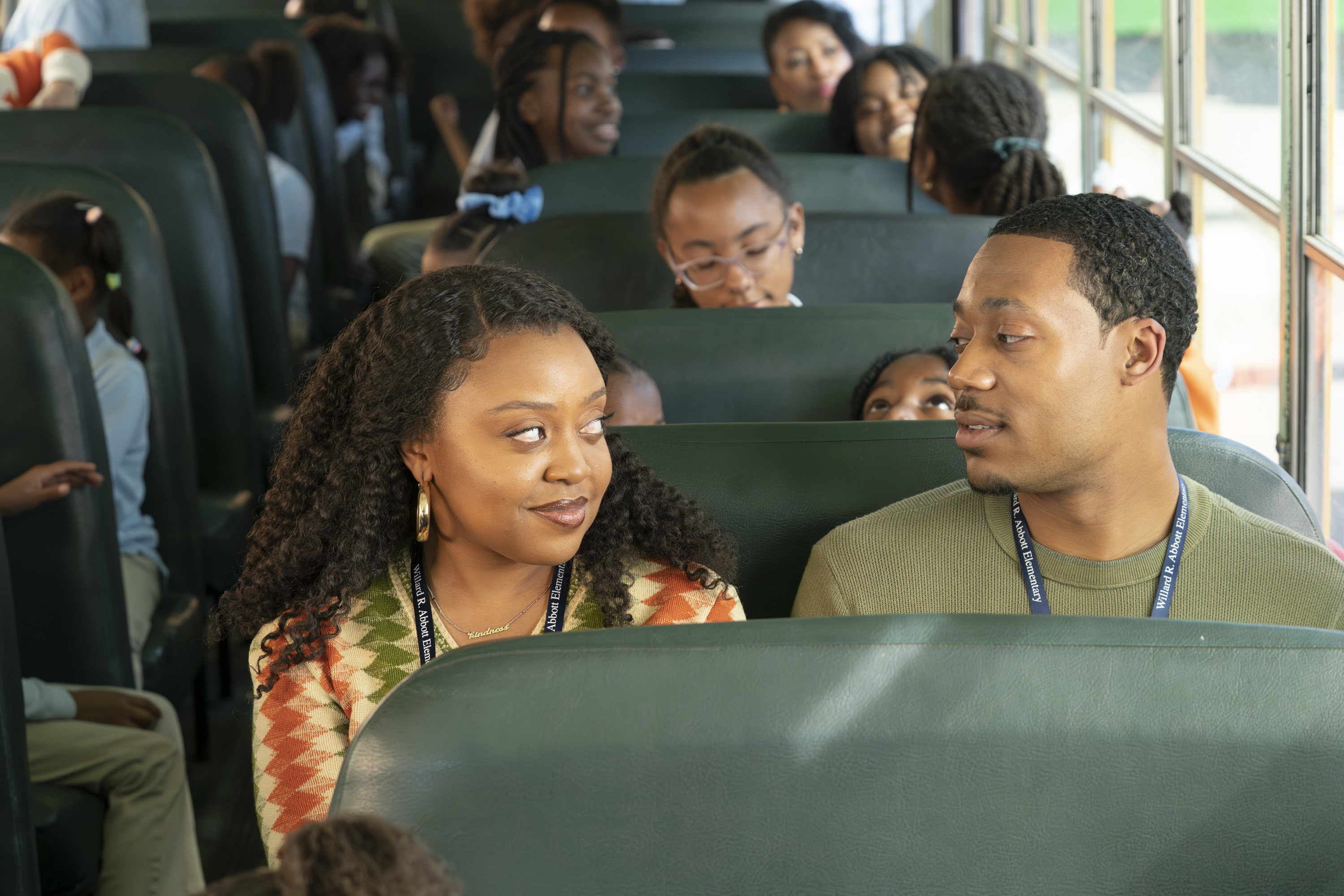 Quinta Brunson and Tyler James Williams sit on a school bus surrounded by children in uniforms, engaging in conversation