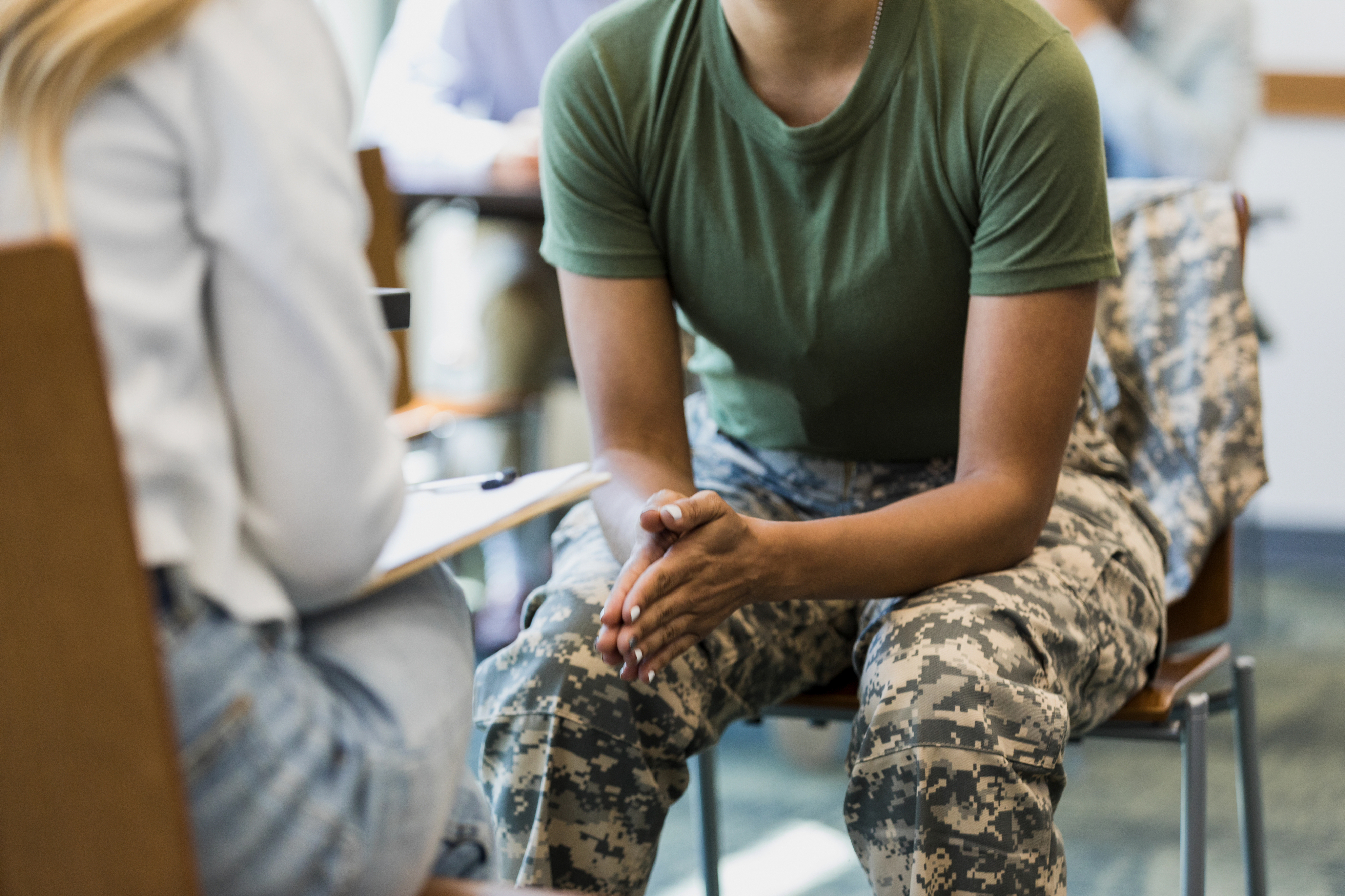 Person in a military uniform sitting and talking with another person who is holding a clipboard in what appears to be an office or counseling session