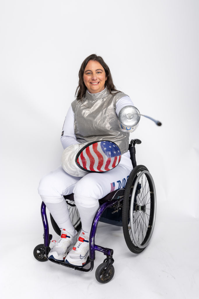 Ellen Geddes in wheelchair fencing gear, holding a sabre and holding a patriotic mask
