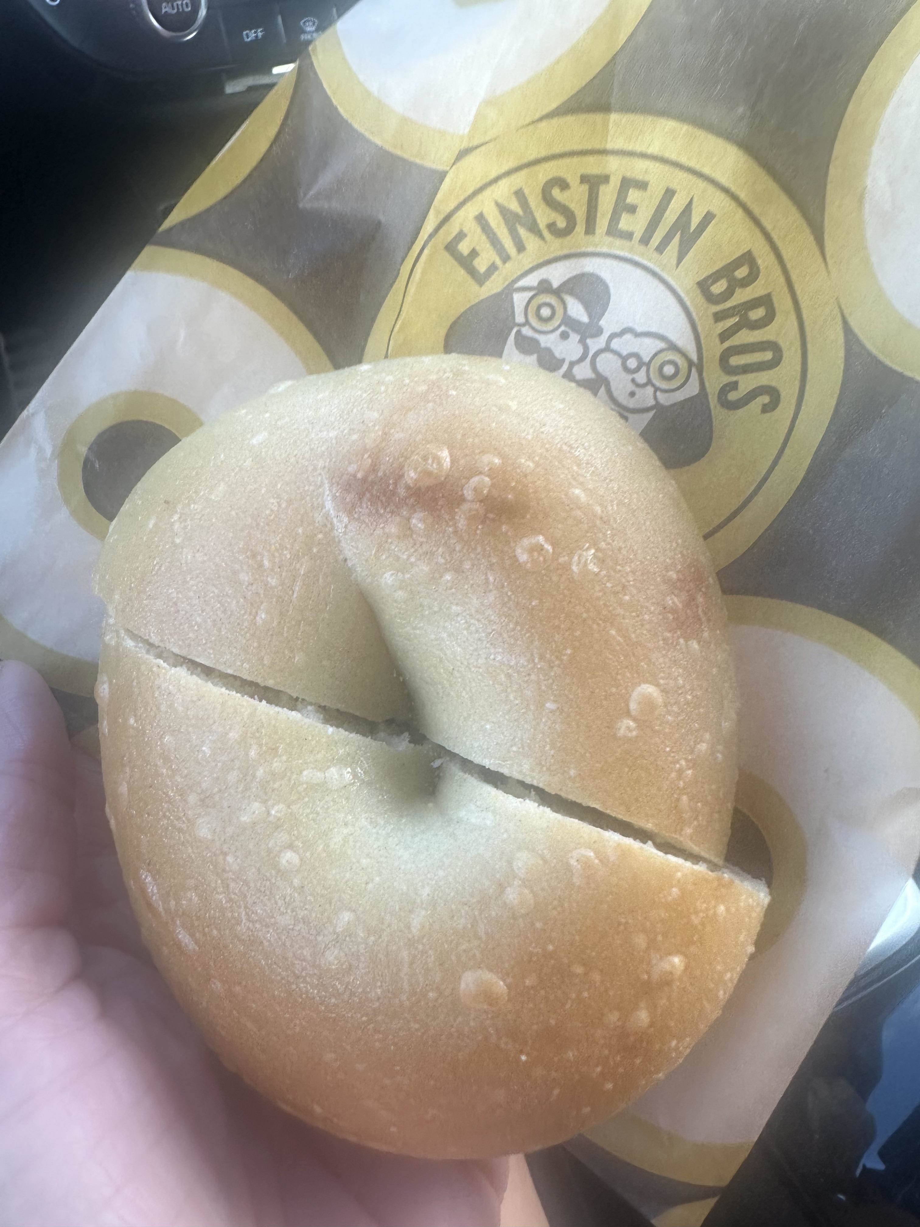 A close-up of a hand holding a bagel from Einstein Bros on a branded paper bag. The bagel is partially sliced