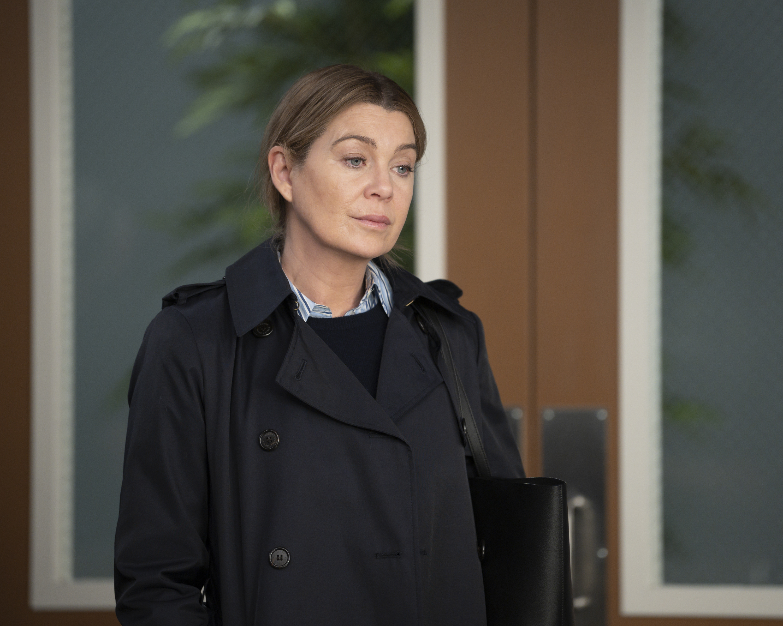 Ellen Pompeo wearing a black trench coat, standing in front of large doors with a contemplative expression