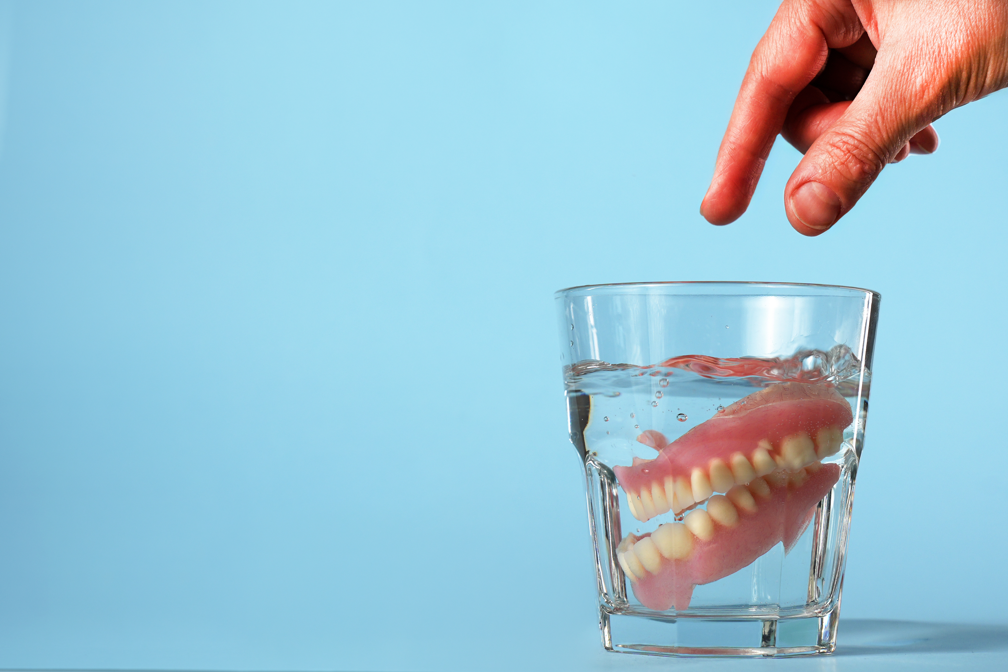 A hand reaches down toward a glass of water with a set of dentures submerged inside