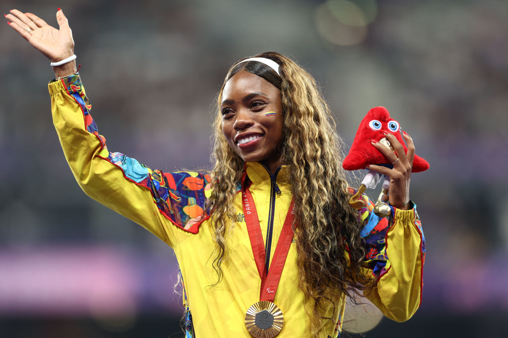 Karen Tatiana Palomeque celebrating with a medal around her neck and holding a plush mascot toy, wearing a colorful jacket with vibrant patterns on the sleeves, smiling and waving, small Colombian flag painted on her cheek