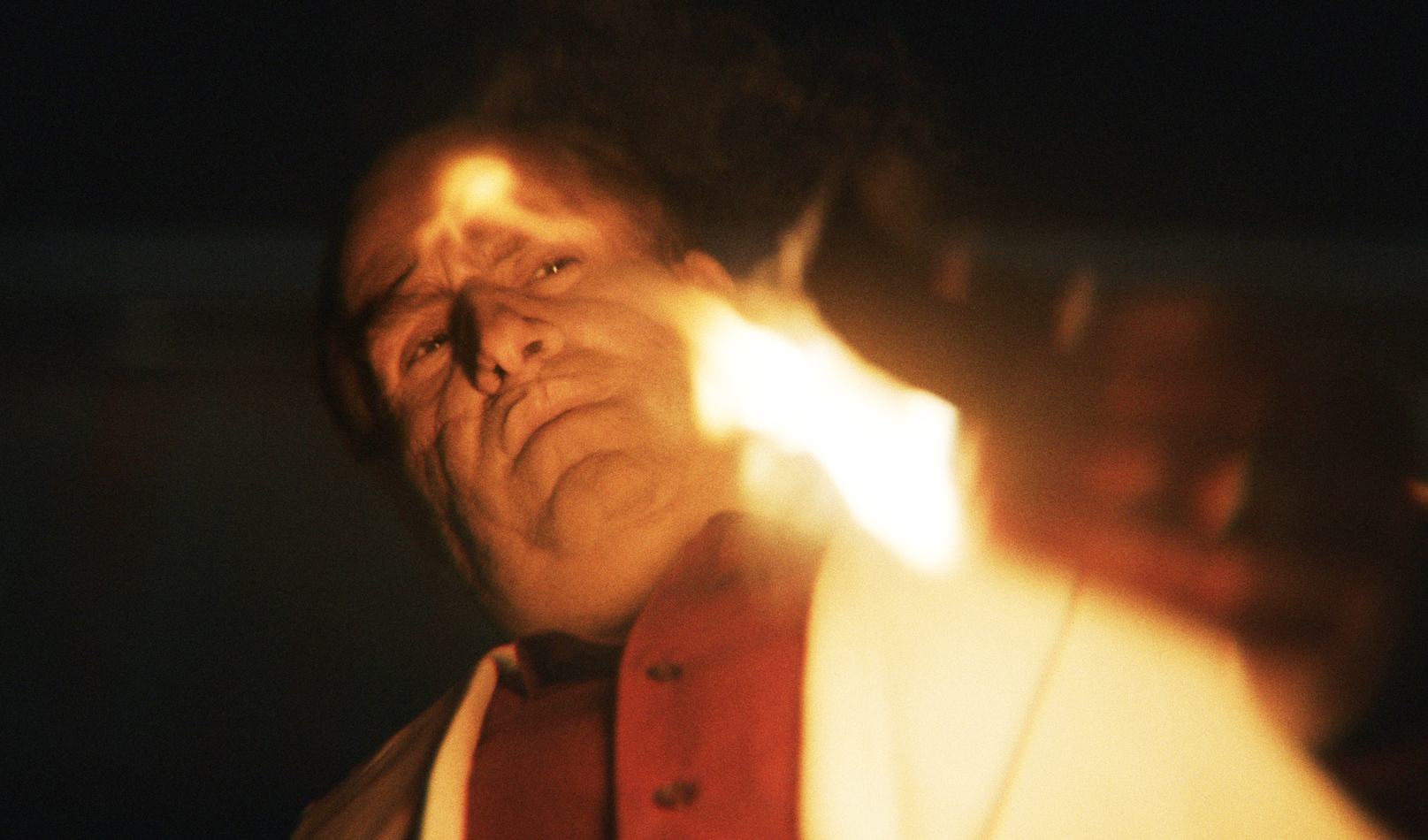 A person in a dramatic scene points a flame towards the viewer, giving an intense and determined expression