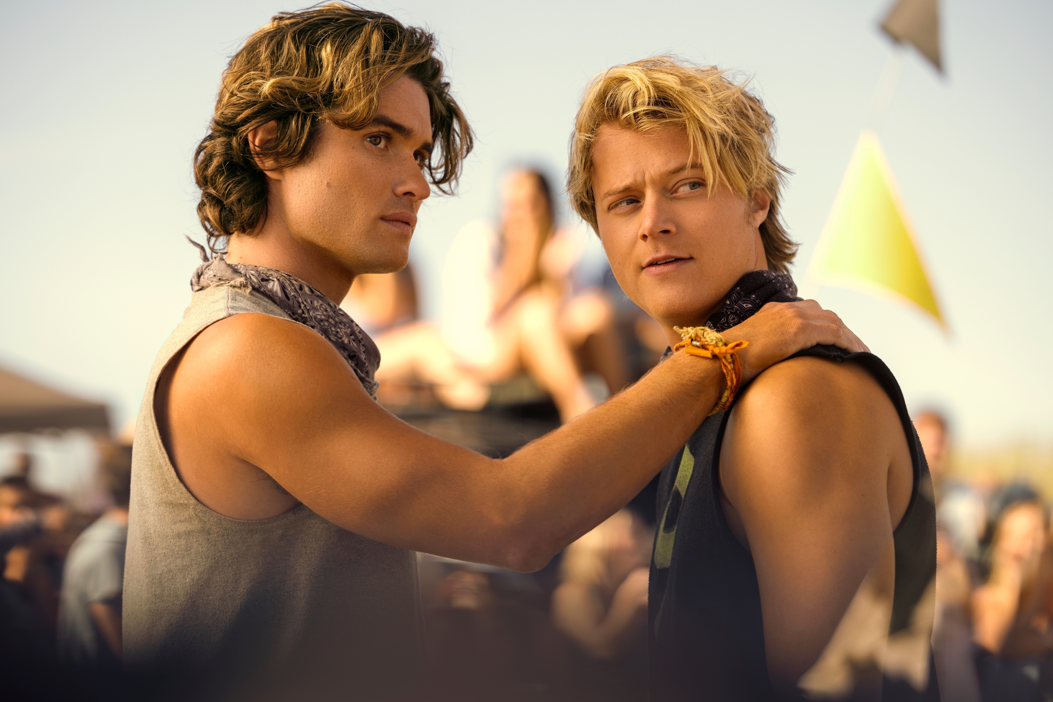 Chase Stokes and Rudy Pankow in casual sleeveless tops, with Stokes placing his hand on Pankow's shoulder, in an outdoor sunny setting